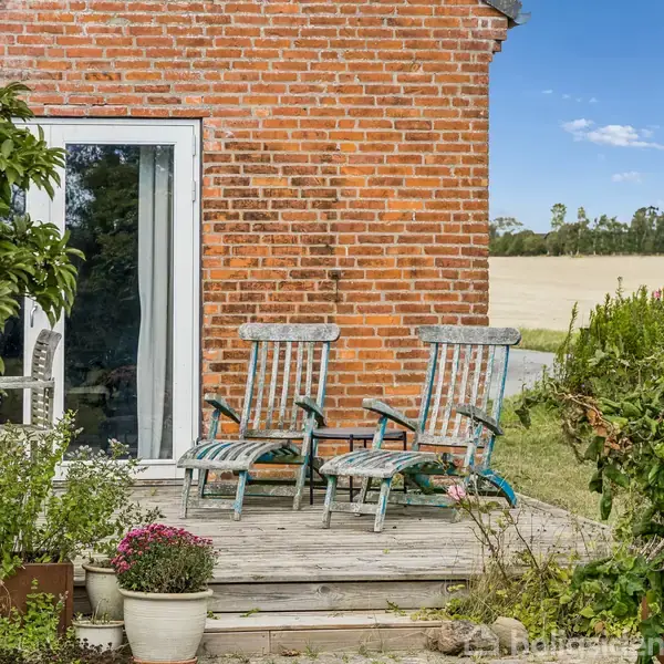 To træstole står på en træterrasse foran et murstenshus. De er omgivet af blomster og planter. I baggrunden ses en mark og et vejskilt med teksten "Strandsandet."