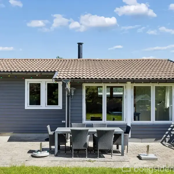 Et sommerhus med mørkt træbeklædte vægge og rødt tegltag. Foran huset står et bord med seks stole på en stenbelagt terrasse omgivet af grøn græsplæne i et solrigt område.