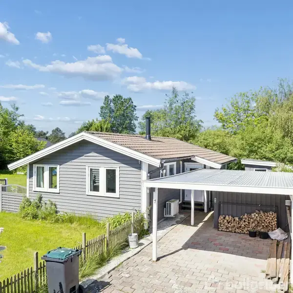 Gråt træhus med carport og brændetække, omgivet af en grøn have med træer og buske under en klar blå himmel. En skraldespand ses foran huset ved en hæk.