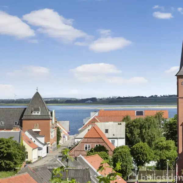 Kirketårn står stille med spidst tag i en byscene. Omkring er røde tegltage og grønne træer. Baggrunden viser en rolig sø under en klar blå himmel med få skyer.