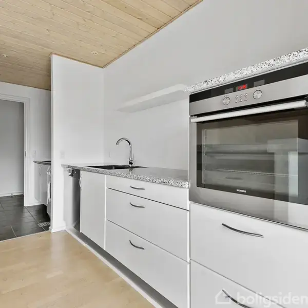 Indbygget ovn i et moderne køkken ved siden af en vask og skuffer, i et rum med lyst trægulv og hvidmalede vægge, set fra en vinkel mod en åben dør.