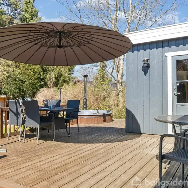 Terrasseområde med stort parasol over et bord med stole. I baggrunden står et spabad omgivet af grønne buske og træer, mens et hus med en dør ses til højre.