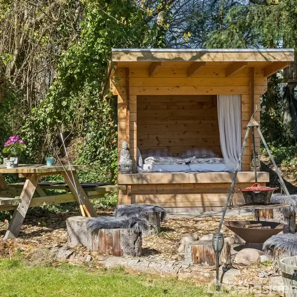 Træhytte med seng indeni står i en have, omgivet af træstubbe og et picnicbord. Haven har grønne planter og blomster, mens træhuset har gardiner trukket til siden.