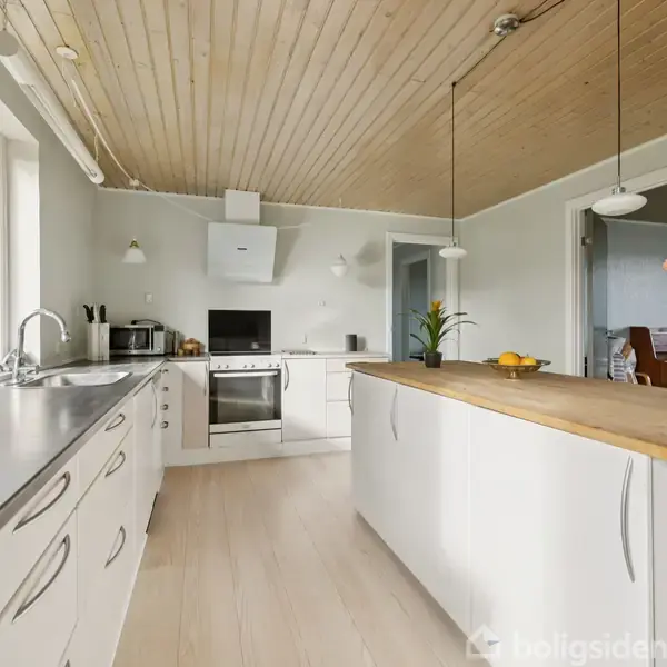 Et moderne køkken med hvide skabe omfatter en central ø. To hængelamper hænger over øen, som rummer en blomsterdekoration. Store vinduer giver lysindfald. Bordet i baggrunden.