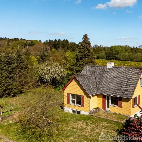 Gult hus står stille omgivet af frodigt grønt landskab og træer under en blå himmel. En rød lade ses til venstre.