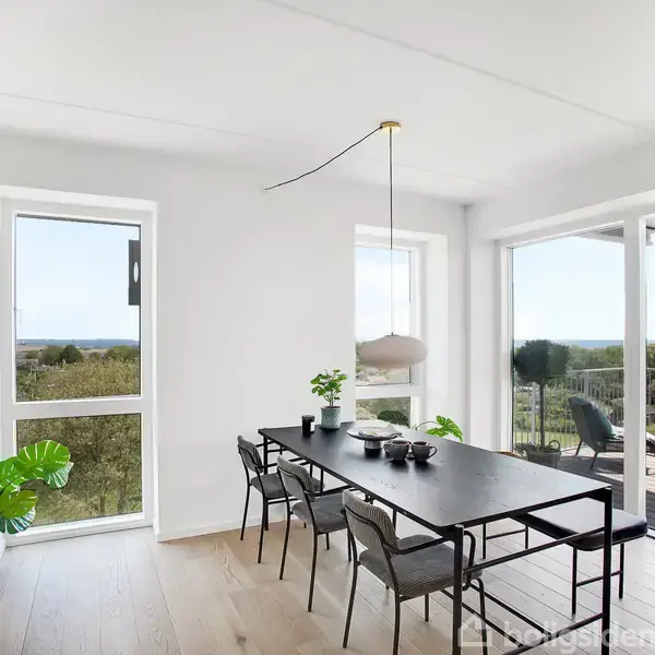 Et sort spisebord med seks stole står i en lys spisestue. Store vinduer i baggrunden giver udsigt til en terrasse og frodigt landskab. En plante og lamper pynter rummet.
