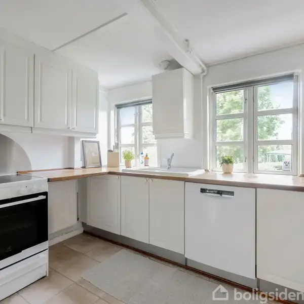 Hvidt køkken med køkkenmaskine og gryde på komfuret. Rummelige skabe omgiver en opvaskemaskine, mens vinduer giver udsigt til grønne træer udenfor.