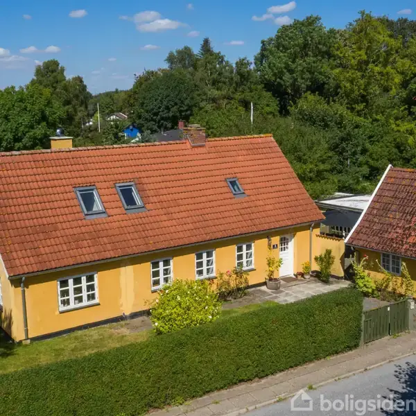 Et gult hus med rødt tegltag og flere vinduer står omgivet af en grøn have og træer. En mindre bygning ligger ved siden af, adskilt af en hæk.