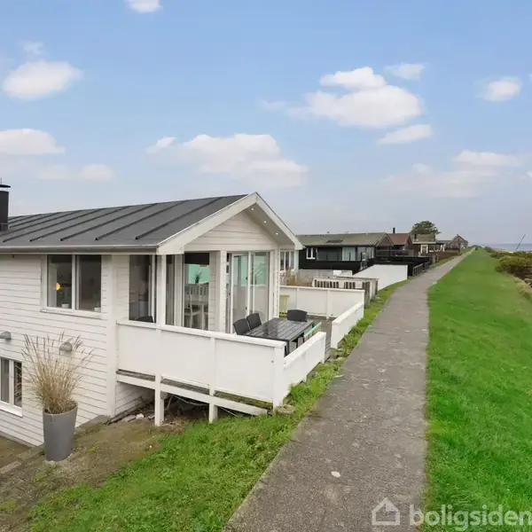 Et hvidt hus står stille ved siden af en havsti; omgivet af grønt græs og en strandbred i horisonten under en blå, skyet himmel.