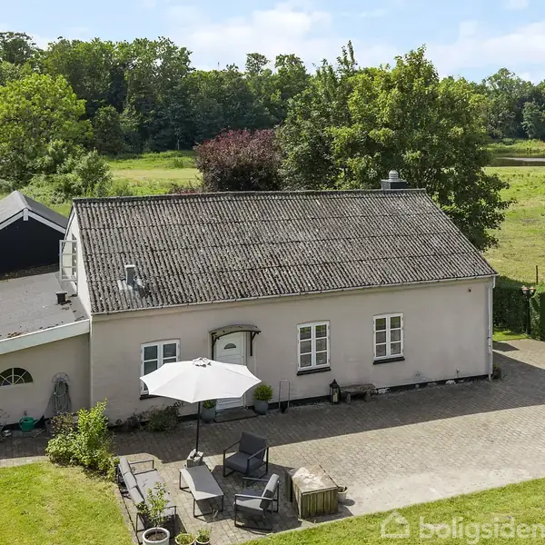 Garderobeområde med jakker hængende på en stang. Åben dør fører til terrasse med grøn udsigt. Til højre er et køkken med ovn. Trægulv og hvidmalede vægge.