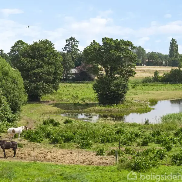 Køkkenø med træskuffer står i midten, omgivet af indbyggede skabe og en håndvask. Rum oplyst af indbyggede loftspotlys. Hvid væg i baggrund med en trappe til venstre.