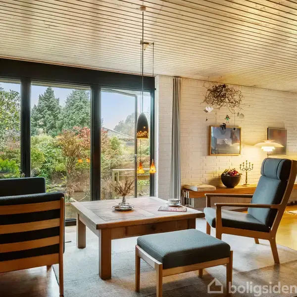 Stue møbleret med træborde og stole, der vender mod store vinduer. Udsigten viser en frodig have. Væggene er hvide mursten, og belysning hænger fra loftet.