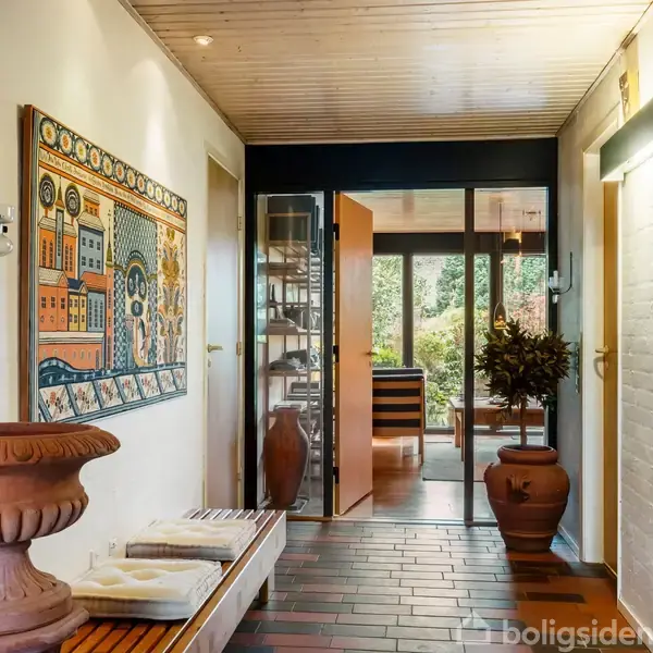 Indretning: En gang med et farverigt vægmaleri, trægulv og loft, fører til en opholdsstue med glasdør og udsigt til en have. Dekorative krukker står langs væggene.