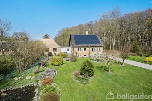 Hus med solpaneler på taget står stille i en frodig have med græsplæne og træer, omkranset af en skov. En lille dam ses i forgrunden.