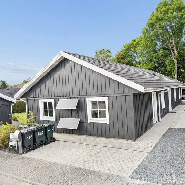 Grå træhus står stille ved siden af indkørsel belagt med fliser; omgivet af grønne buske og træer. To affaldsbeholdere med numre "66" står foran.