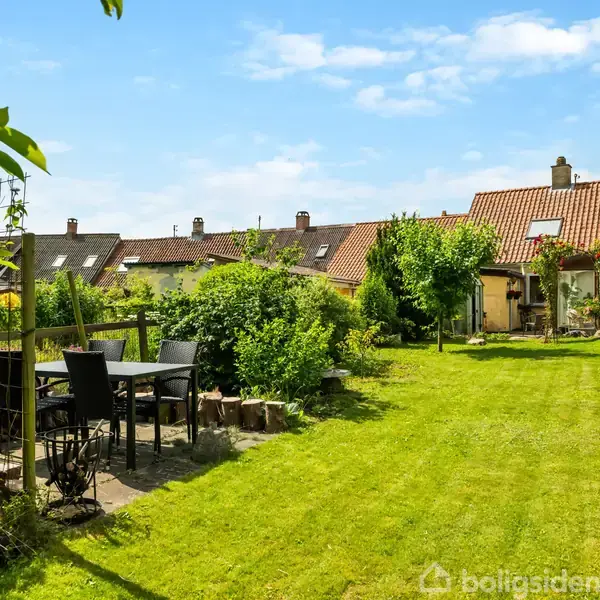 Havemøbler står på en terrasse ved siden af en velplejet græsplæne, omgivet af buske og små træer. I baggrunden er der flere rækkehuse med røde tegltage.
