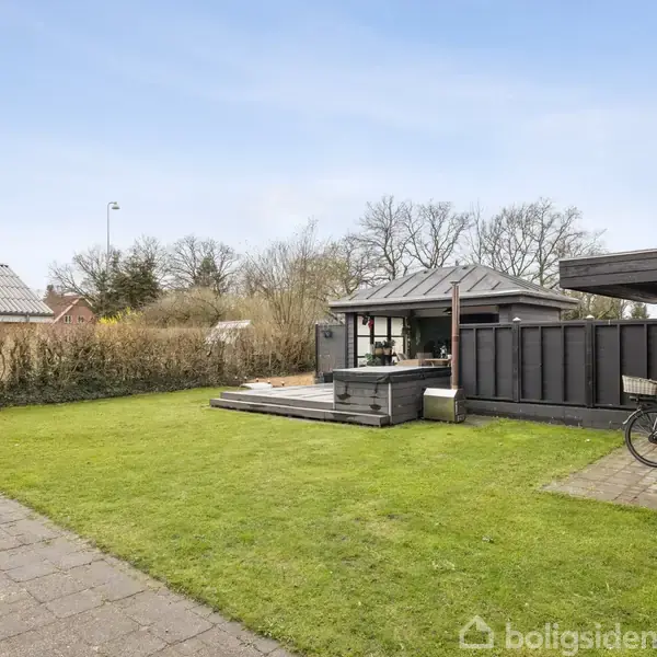 En velplejet græsplæne med flisebelægning i forgrunden leder til et lille sort skur med tilhørende terrasse. I baggrunden ses en hæk og nogle træer.