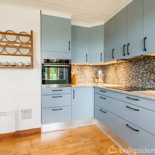Blåt køkken med indbyggede skabe og skuffer. Bagvæggen er dækket af mosaikfliser, og der er en dobbelt ovn indbygget. En hylde til kopper hænger på væggen. Parketgulv i træ.