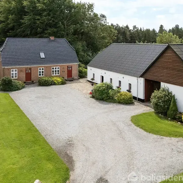 To huse med sort tag står ved en grusvej omgivet af grønne buske og plæner. Flagstang og havebænk ses i nærheden. Træer danner baggrund. Ingen synlig tekst.