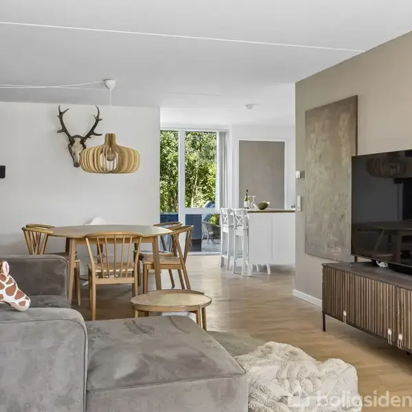 Stor stue med en grå sofa, hvor en giraf-pude ligger. Et tv står på et træskab, og der er et spisebord med stole. En køkkenø ses i baggrunden.