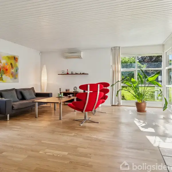 Stue med grå sofa og rød lænestol omkring et træbord, stor lysende gulvlampe og farverigt maleri på væggen. Store vinduer åbner mod grøn have. Parketgulv.