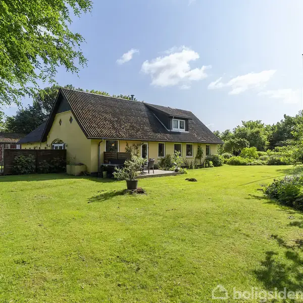 Et gult hus med rødt tag står stille i en frodig grøn have. Træer og buske omkranser huset, mens en klar blå himmel ses ovenfor.