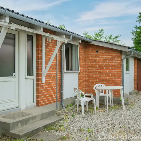 Rødt murstenshus med hvid indgangsdør og vinduer. Foran huset står et hvidt plastikbord med to stole på en grusbelagt gårdsplads. Grønne buske omkranser området under en klar himmel.