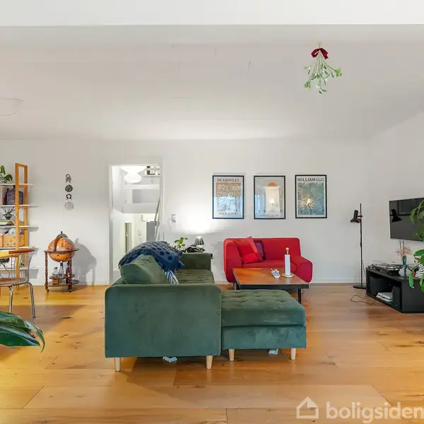 Stue med grøn sofa og sofabord, rød sofa mod hvid væg med tre plakater, træhylde med planter til venstre; hjørne med globusbar og reol. Trægulv og planter skaber en hyggelig atmosfære.