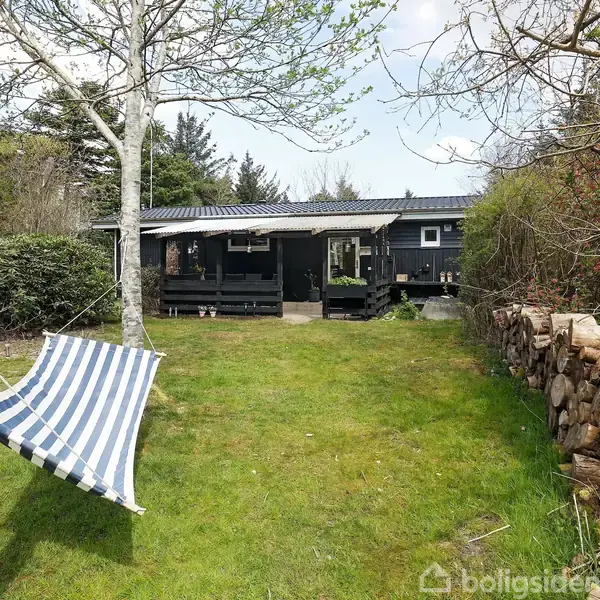 En hængekøje af blå og hvide striber hænger fra et træ i en have foran et sort træhus. Græs, træer og en brændestabel omgiver scenen.