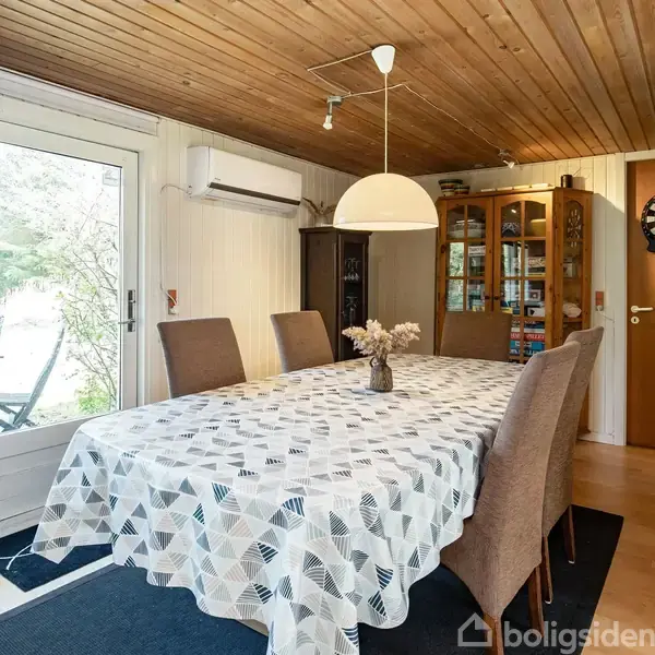 Spisebord med dug og blomster i vase. Seks stole omkring bordet. Indendørs i et rum med trægulv og træloft. Glasvitrine, skabsdør og dartskive i baggrunden.