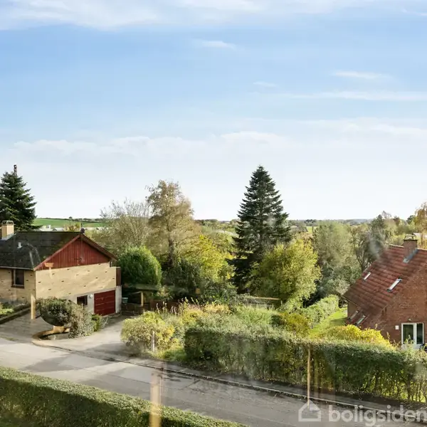 To huse står stille på en stille villavej, omgivet af træer og buske i et naturskønt landskab under en blå himmel.