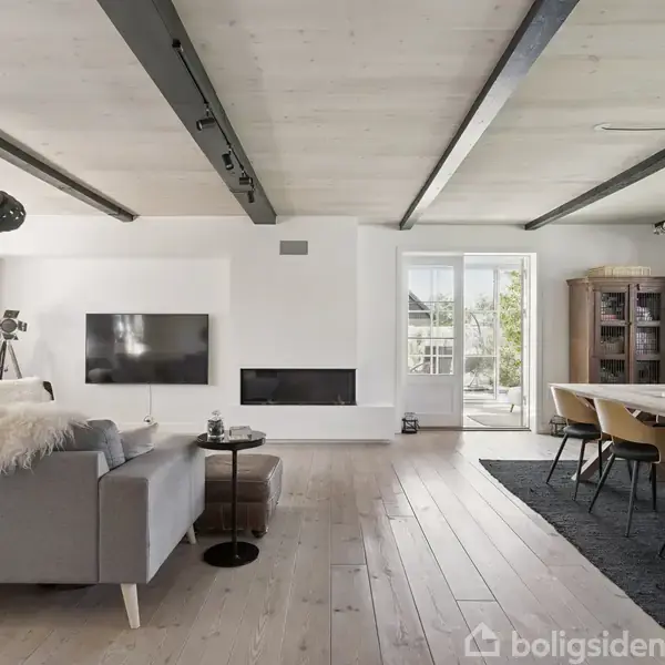 Stue med gråt sofaarrangement ved siden af moderne fladskærms-TV og indbygget pejs. Spisebord med stole til højre. Lyse trægulve og loftsbjælker supplerer de store vinduer med naturligt lys.