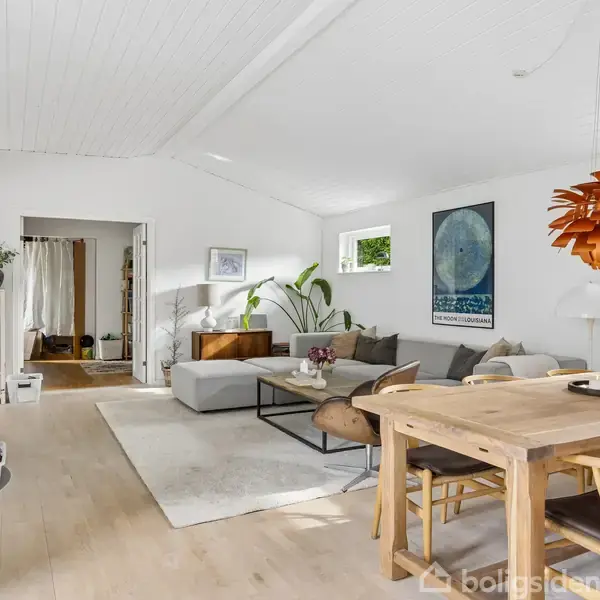 Stue med en grå sofa og sofabord, placeret ved siden af en brændeovn. Et spisebord med stole står i forgrunden. Væggene er hvide med moderne plakater.