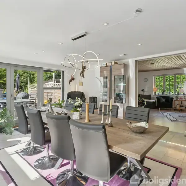 Langt spisebord med sorte stole står i et lyst rum med glasdøre til terrasse. Der er moderne møbler og grønne planter. Rummet åbner op til en stue med sofa.