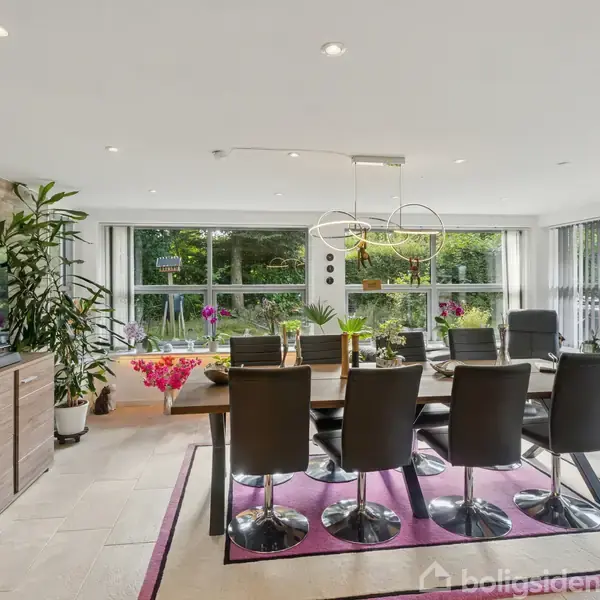 Spisebord med otte sorte stole står i et lyst rum med store vinduer, planter og udsigt til haven. Omkring bordet hænger en moderne lampe.