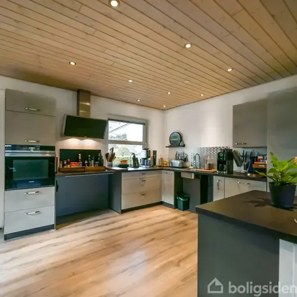 Køkken med moderne grå skabe og træloft, indrettet med apparater og køkkenredskaber. Et stort vindue giver lys, og der står stearinlys og en plante på køkkenbordet.