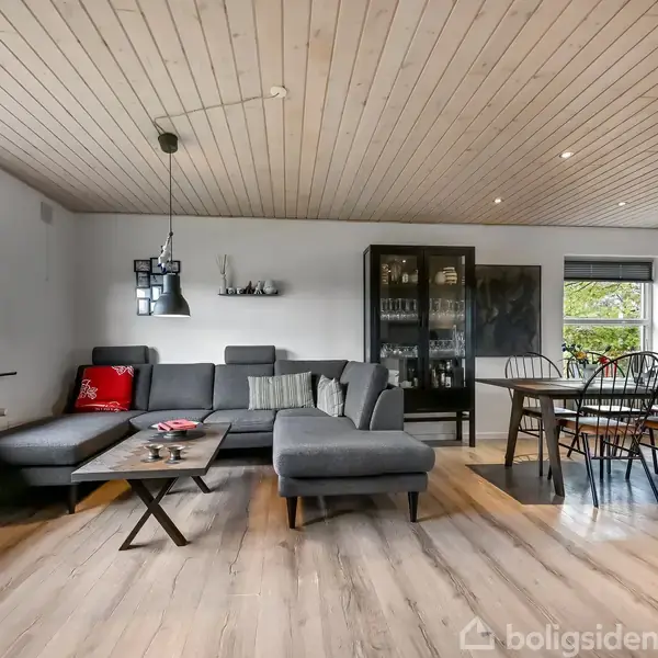 En grå sofa med puder står i en lys stue. Ved siden af er et spisebord med stole. To vinduer giver udsigt til træerne udenfor. En sort loftlampe hænger over sofabordet.