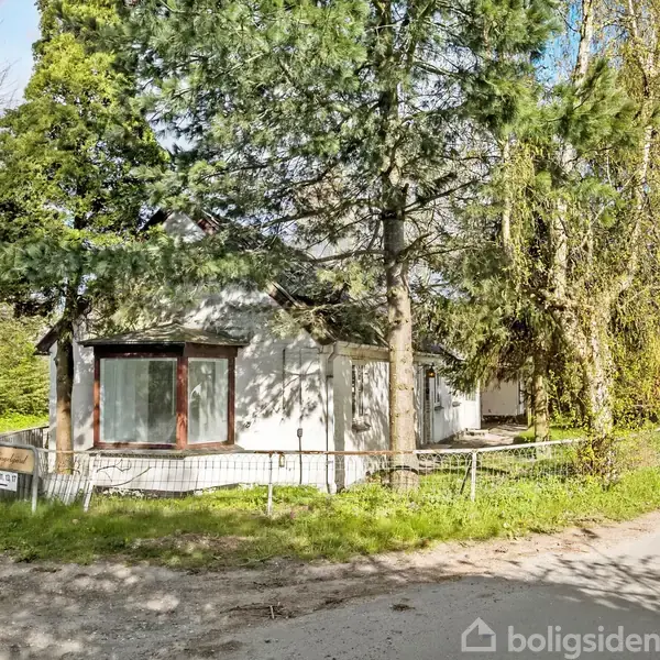 En lille hvid bygning står blandt høje træer ved en vej. Skiltet ved vejen viser "Åbakken". Området omkring bygningen har grus og grøn vegetation.