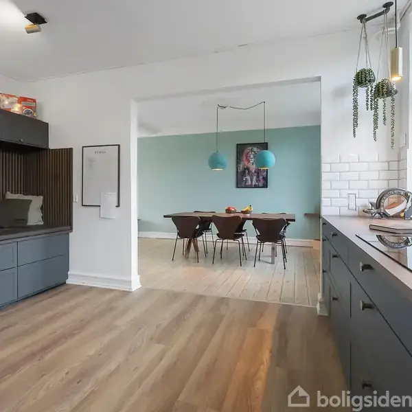 Køkken med grå skabe og træbordplade. Udsigt til spisestue med træbord, seks stole og blå væg. Loftbelysning over spisebordet, planter hænger fra loftet, store vinduer til højre.