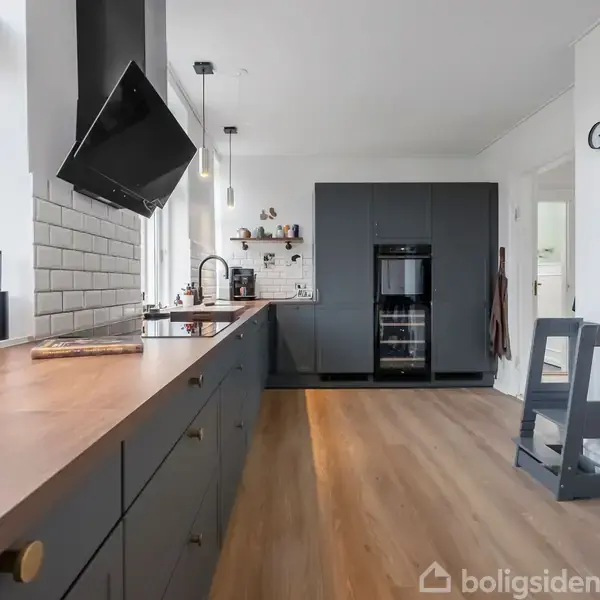 En moderne køkkenindretning med mørke skabe og skuffer, og en træbordplade. En stor emhætte hænger over en kogeplade. Rummet er lyst med vinduer og stilfuld belysning.