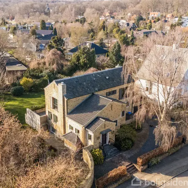 En stor, gul murstensvilla står stille i et villakvarter omgivet af træer og hække. Den ligger i en rolig gade med flere huse i baggrundens landskab.
