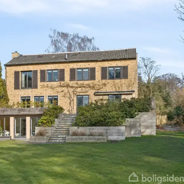 Toetagers murstenshus står i en grøn have med træer og buske. En trappe forbinder husets niveauer. Skyfri blå himmel i baggrunden flankerer huset.