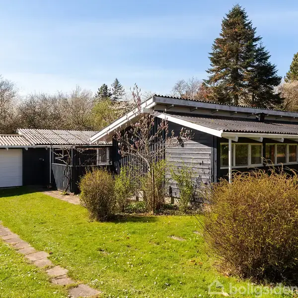 Et sortmalet hus med fladt tag står i en frodig have. Huset har store vinduer og ligger ved siden af en garage. Omkring huset ses høje træer.
