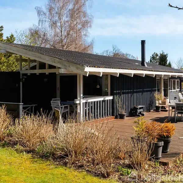 En sortmalet træhus med hvidt tag har en overdækket terrasse med møbler. Det står i en have med græsplæne og blomsterbeplantning, omgivet af træer under en blå himmel.