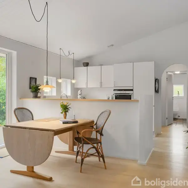 Et lyst køkkenalrum med åben layout; spisebord med to stole ved siden af en køkkenø. Store glasdøre leder ud til haven; hvide vægge og træplankegulv.