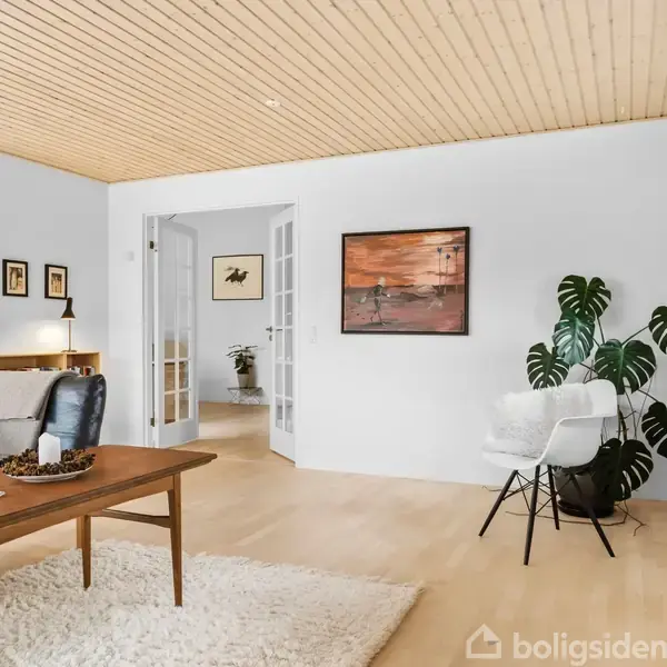 En stue med sort lædersofa og træbord står på et lyst gulvtæppe. En stol og plante er placeret ved væggen med kunst og en fransk dør.