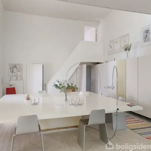 Et stort, lyst rum med en stor hvid bord, omgivet af moderne stole. Bagerst ses en trappe, hvidmalede vægge, minimalistiske dekorationer og en farverig løber på gulvet.