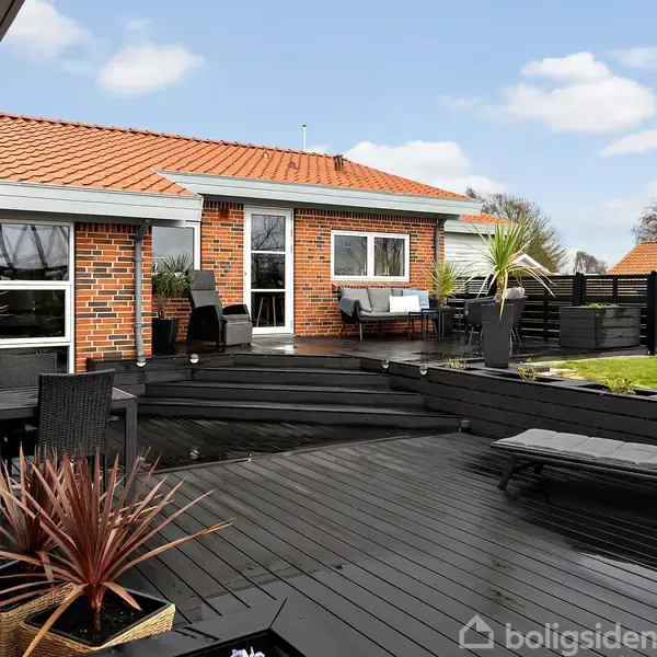 Rød murstenshus med udendørs terrasse, hvor sorte trægulve og moderne havemøbler ses. Terrasse omkranset af plads til afslapning. Omkringliggende græsplæne og klar himmel i baggrunden.