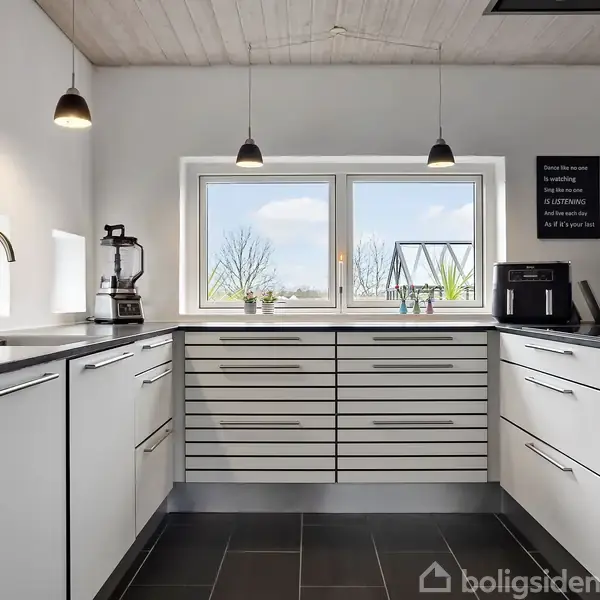 Et moderne køkken med hvide skabe og sorte bordplader, der er velorganiseret med en blender og kaffemaskine. Store vinduer giver udsigt til en have med en gynge. På væggen står der: "There is nothing that a good meal cannot cure" i sorte og hvide nuancer.
