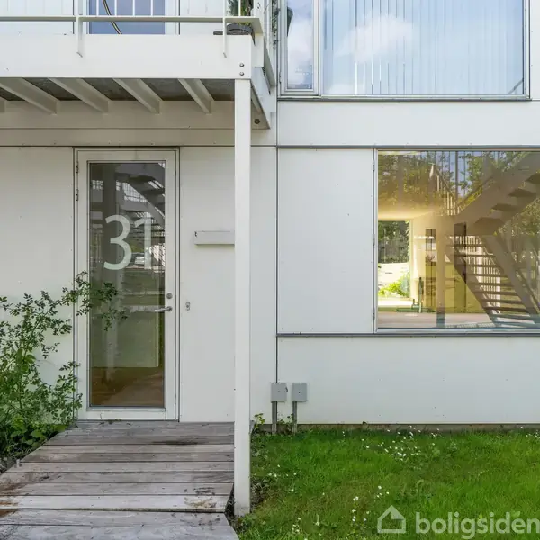 Dør med tallet "31" i glas, stående i en moderne, hvid bygning. Trægang fører til døren, omgivet af grønne planter og græs. Store vinduer afslører indvendige trapper.